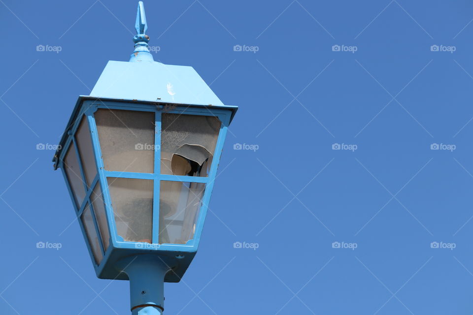 Old blue lantern lamp stand light pole with broken glass against vivid clear blue sky 
