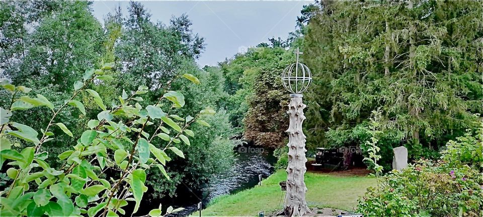 A lush park like garden extends to border a small stream somewhere in „Mecklenburg“. and art object adorns the lawn. 2022. Hypnotic Productions