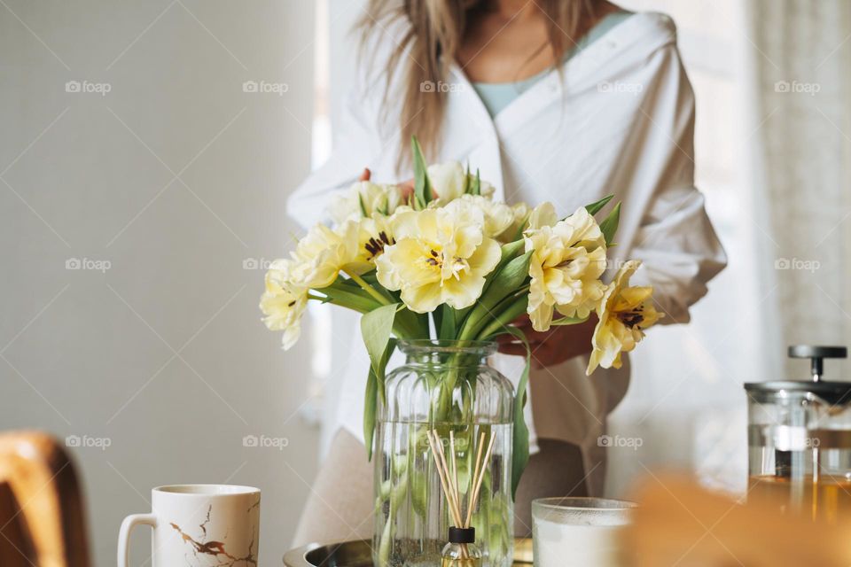 Young beautiful smiling woman forty year with blonde long hair in white shirt with bouquet of yellow flowers in hands near dinner table in bright interior at the home