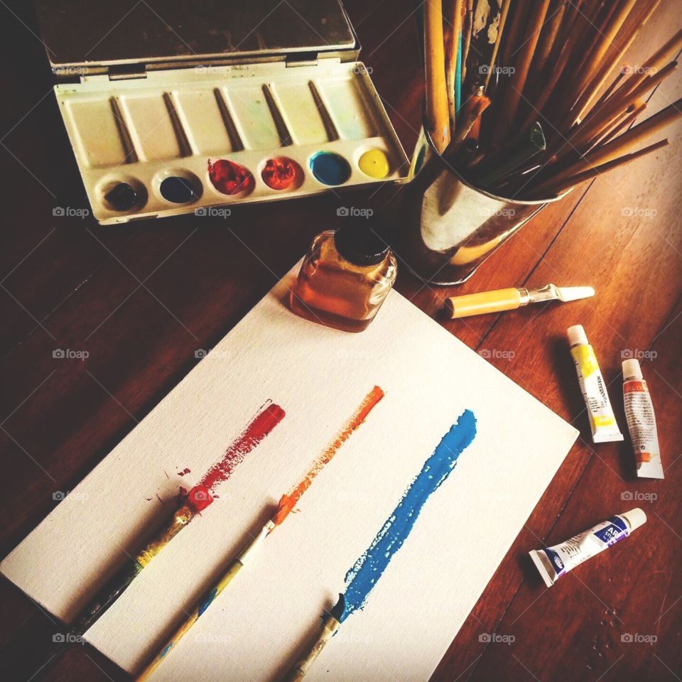 A white canvas with simple, multi-colored brush strokes on it surrounded by paints and painting tools on wooden surface.