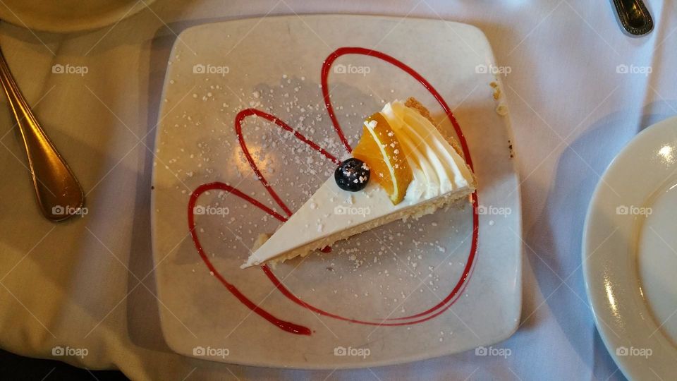 a yuzu lemon cheesecake slice with yuzu wedge and blueberry garnish, strawberry syrup drizzled in a tulip shape and powdered sugar sprinkled across the white ceramic plate.