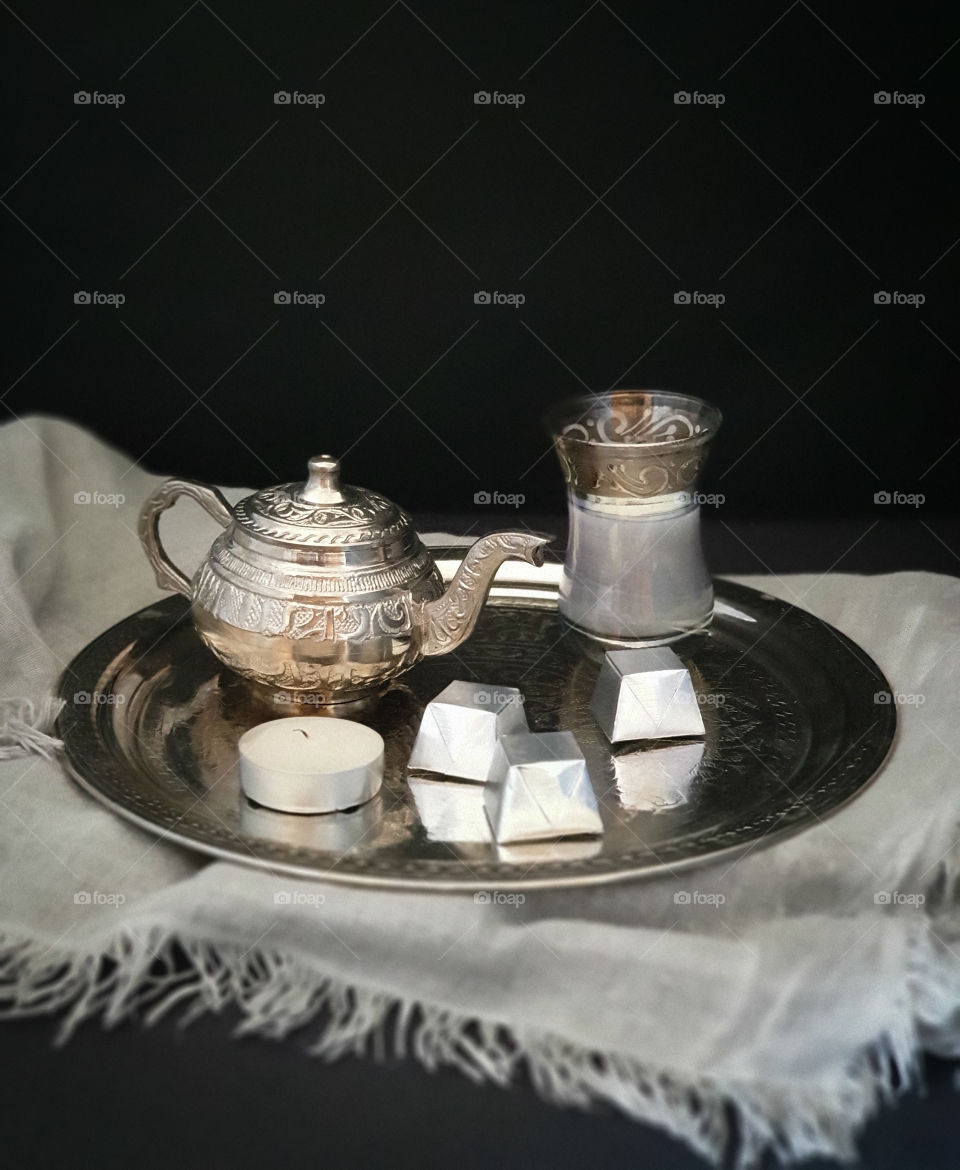 Dark oriental still life.  Silver ornamental tray with a small silver teapot and a glass with tea with milk, sweets and a candle on gray beautiful textile.  Tasty snack