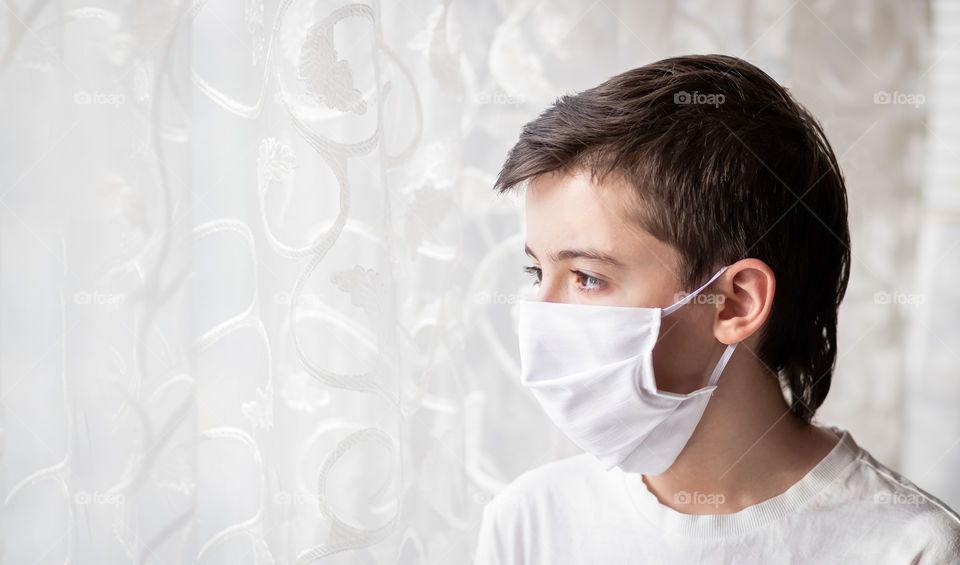 Portrait of child in a protective white mask near the window. House pandemic period. Concept of the fight against coronavirus covid-19.