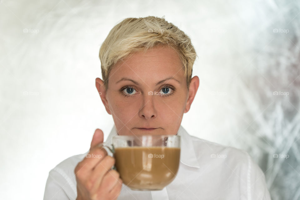 woman in white clothes, with short blond hair loock straight in camera. she is holding glass cup of  coffee