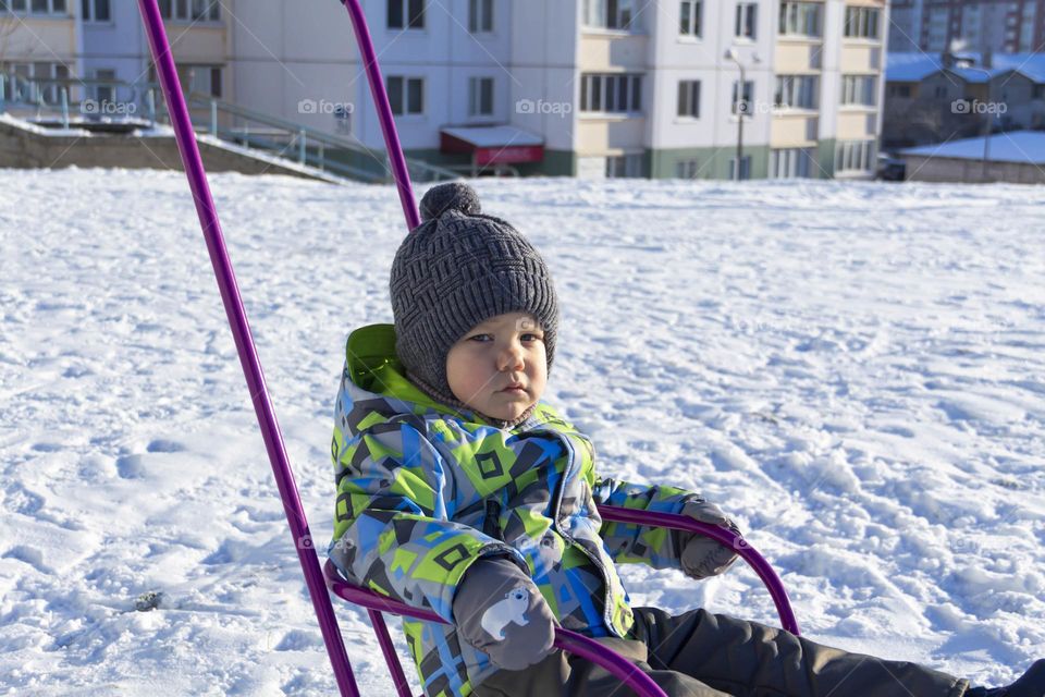 A child with a serious expression on his face in winter clothes jackets, pants, hat and boots in winter on white snow on the street and in the park in nature sledding and playing winter fun.