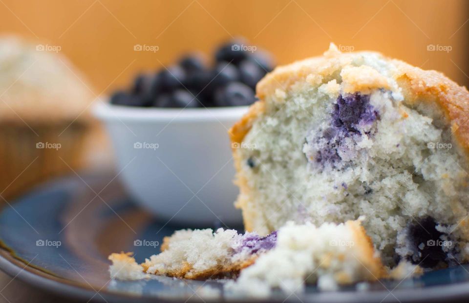 blueberry muffin broken open to show white inside of muffin speckled with purple colors of baked blueberries on a blue plate with a blurred white bowl full of blueberries and an orange background