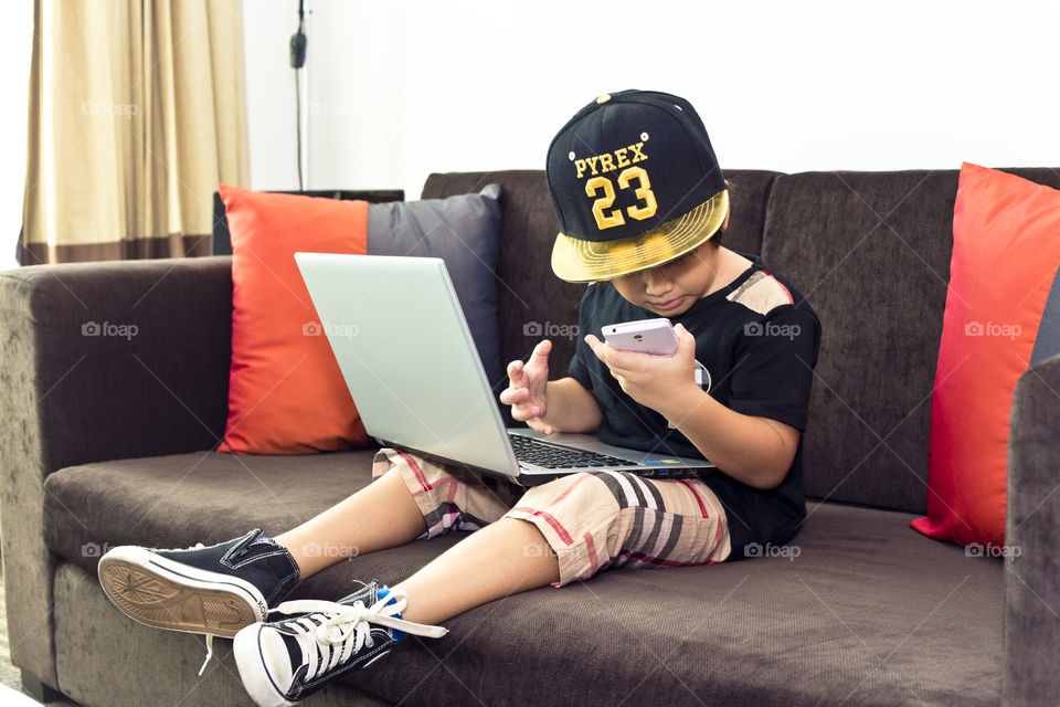 Kid and technology. Young boy Thithi holding a mobile phone while working on his laptop