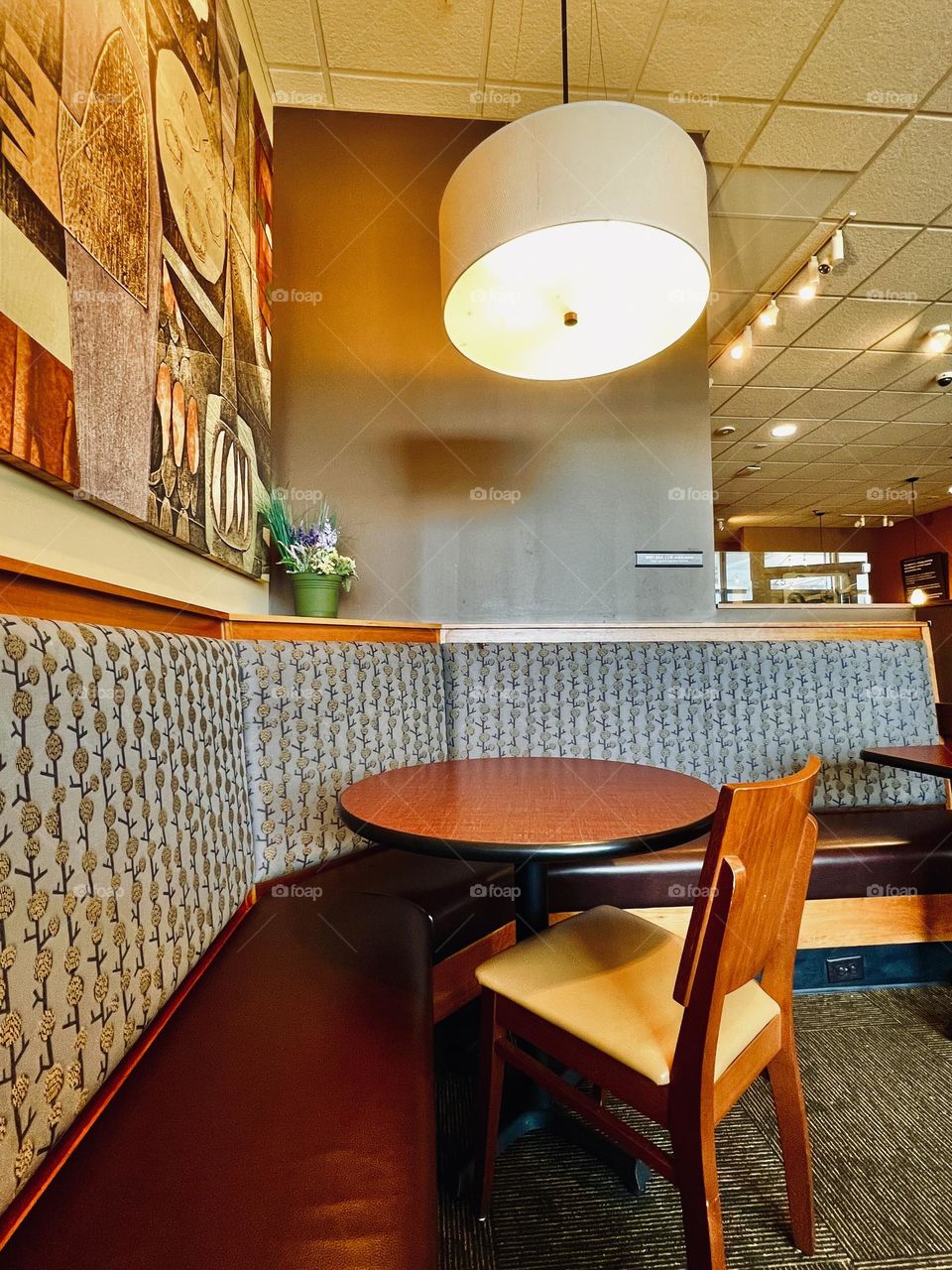 Corner banquette seating in coffee shop. Round wooden table and chair against padded bench with round  drop light fixture. Abstract art  and small potted plant with neutral warm color palette.