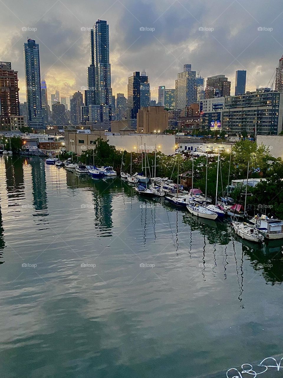 This is a twilight time view from the “Pulaski Bridge” at “Newtown Creek” in LIC, Queens onto various boats by the shore in the waters of the “East River” that shimmers in various shades of a deep metallic silver tonight. 2023. Hypnotic Productions