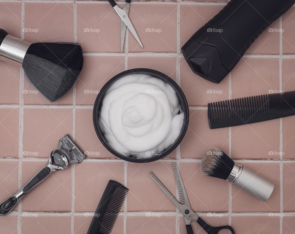 Porcelain bowl with shaving foam, and around it are brushes with natural bristles for shaving, combs, scissors, a bottle of aftershave cream and a razor in the center on a brown natural tiled background, flat lay close-up. The concept of a man barber