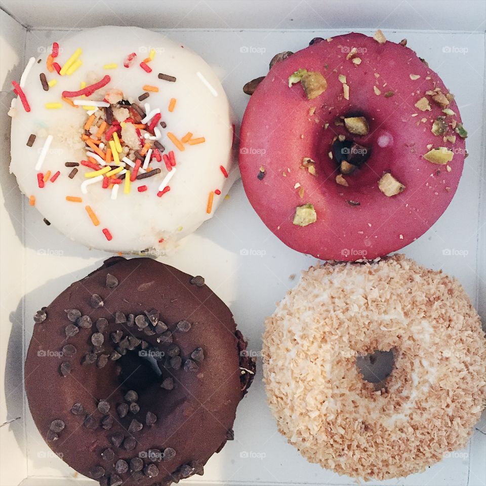 An assortment of four donuts, featuring chocolate, coconut, strawberry and vanilla flavours, topped with crumbled nuts, sprinkles, coconut flakes and chocolate chips. 