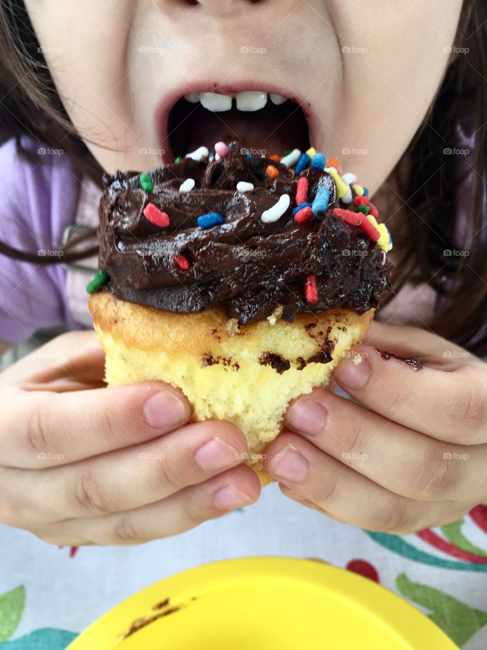 Little girl taking a bite out of a vanilla cupcake with chocolate frosting and rainbow sprinkles 
