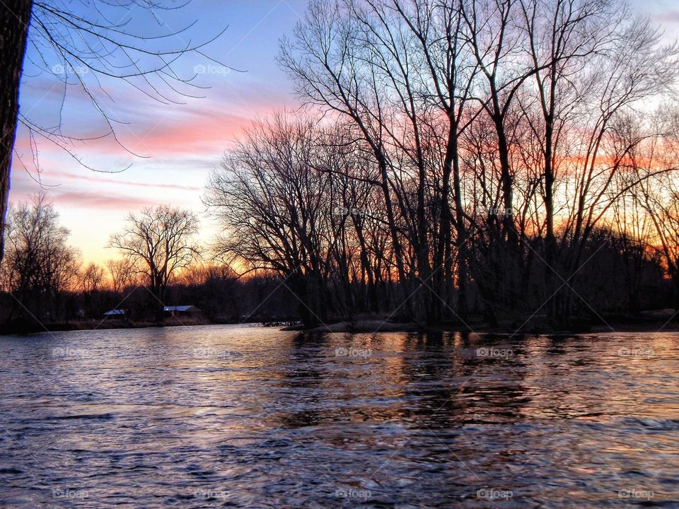 Sunset over Michigan river creating pink orange and purple clouds in the blue sky and casting a reflection of the colors on the water. The trees create a shadow silhouette against the bright sky