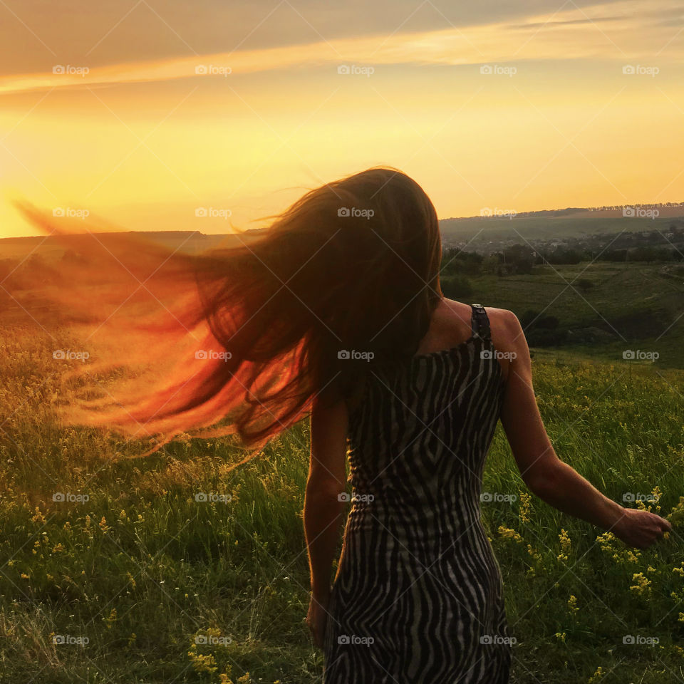 Young woman catching sunset sunlight by her long hair in the wind at the summer field 