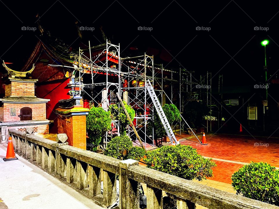 A temple under construction. It is nighttime, and lots of metal ladders are propped against the red temple. 