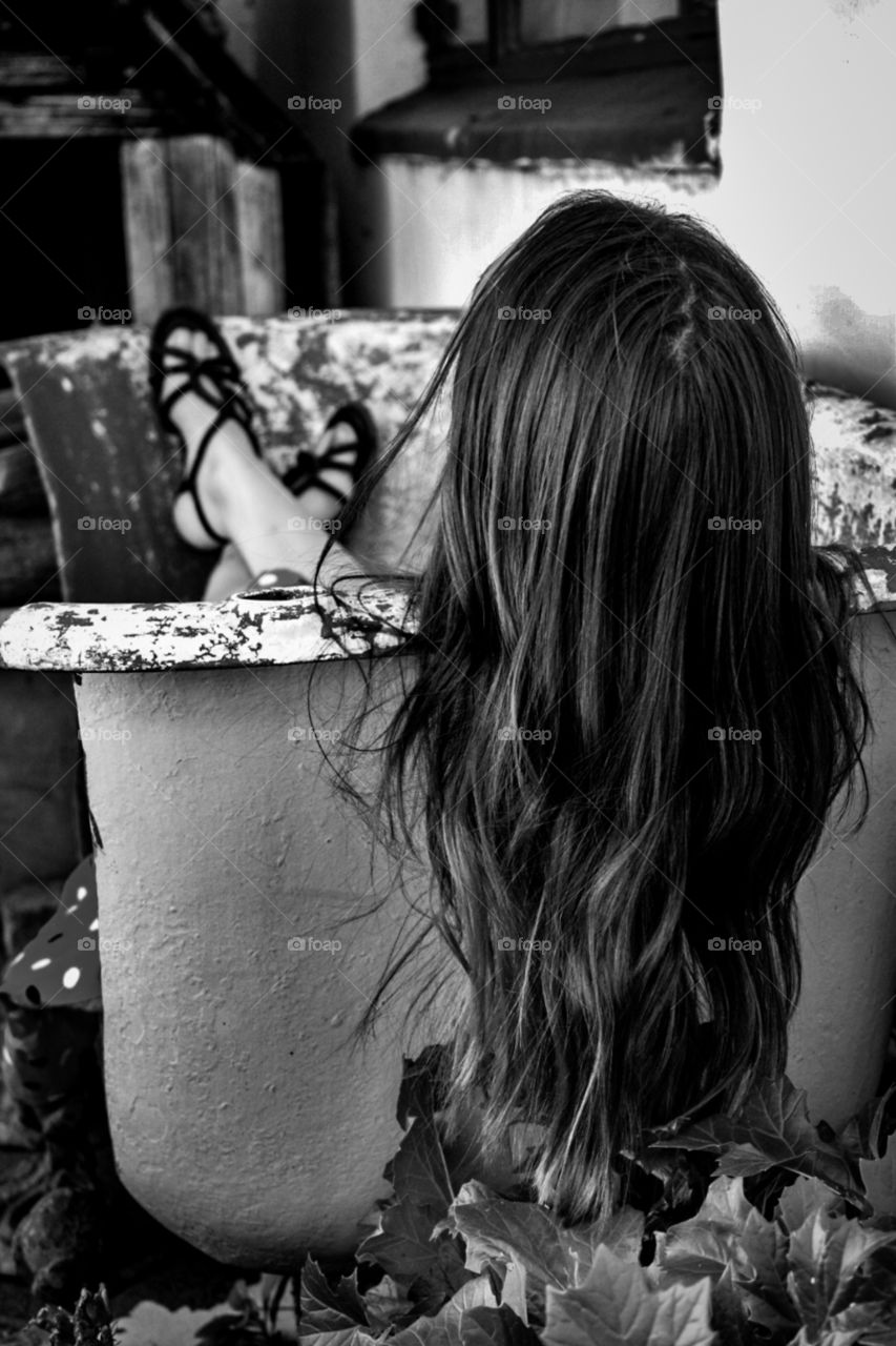 a women laying in a old bathtub that was cut in half, where you can only see the back of her head with long hair hanging down
