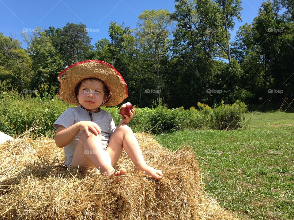 A toddler boy sitting on a hay stack in the sun and eating a fresh picked apple.