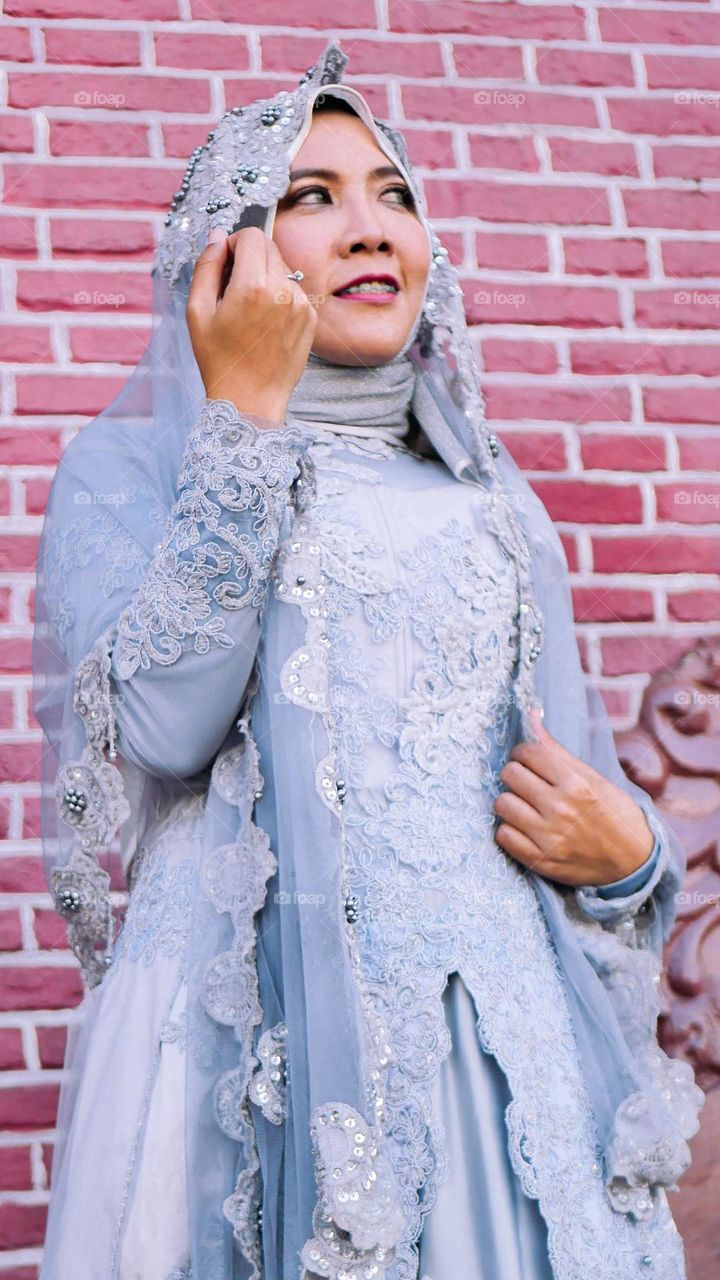 Portrait of young and beautiful Asian muslim woman wearing silver robe with matching hijab and headscarf, holding headscarf and smiling, Red brick wall background. for the wedding.