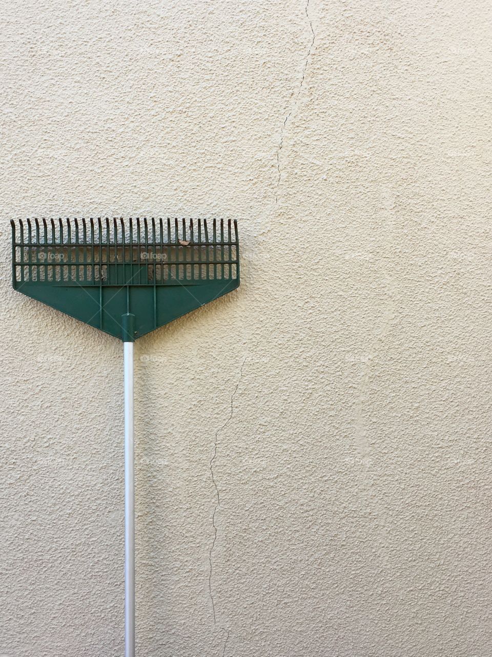 Garden rake leaning up against old exterior stucco wall which has a crack running through it. Rake to left of image, negative space minimalist image