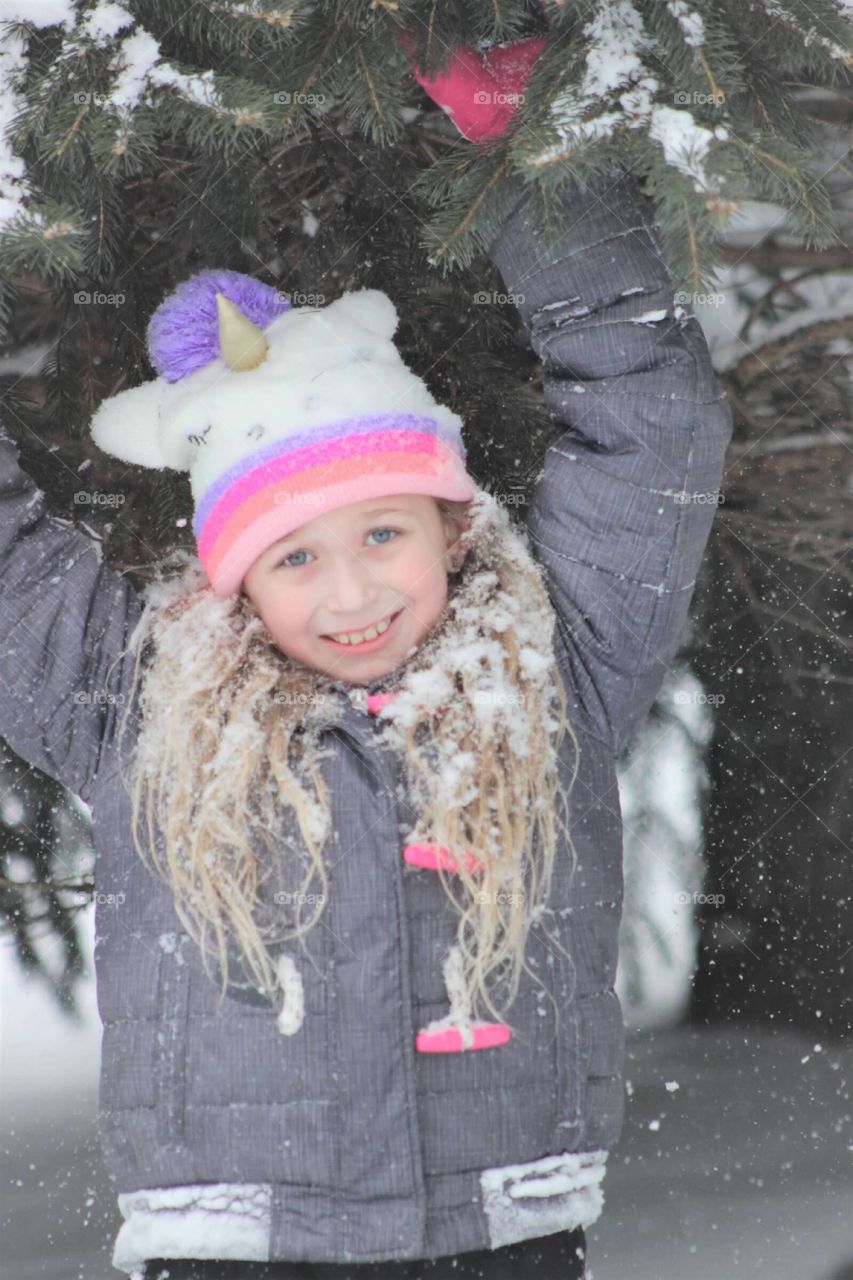 Pretty little girl in a unicorn hat getting covered with snow as she plays underneath a snow covered Pine. 