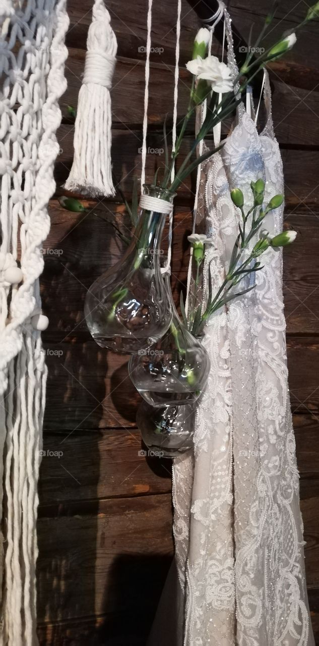 The wedding dress is hanging next to the macrame and the glass bottles with the white carnations.