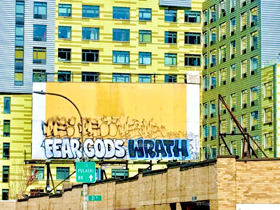 “FEAR GOD’S WRATH” says the graffiti inscription on the bright yellow construction site wall in front of the also bright yellow building in Long Island City, Queens, NY photographed on a sunny afternoon in the summer of 2021. Hypnotic Productions