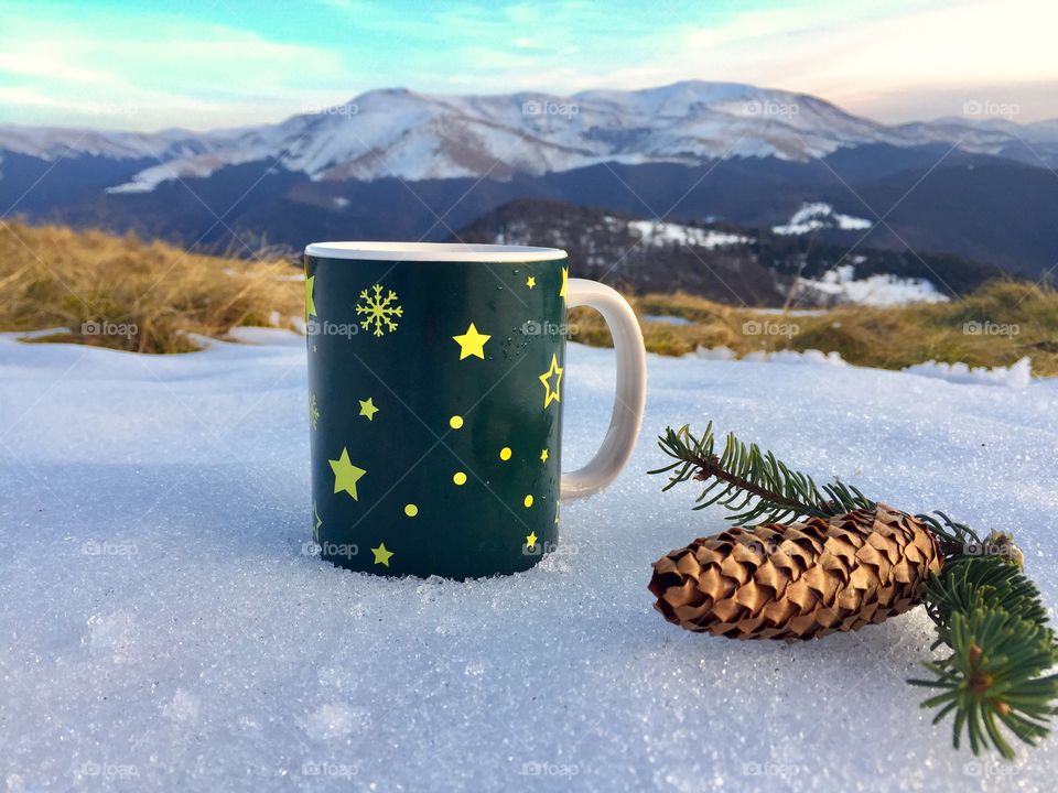 Snowflakes Christmas mug placed on snow with pine cone and evegreen branches beside and snowy mountains in the background