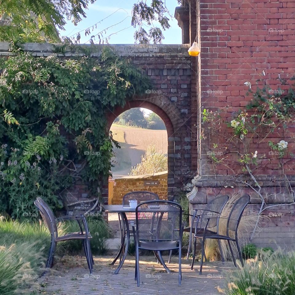 Early autumn U.K empty table and chairs outside with planting around. Arch doorway showing rolling sunny golden fields through it in the background.
