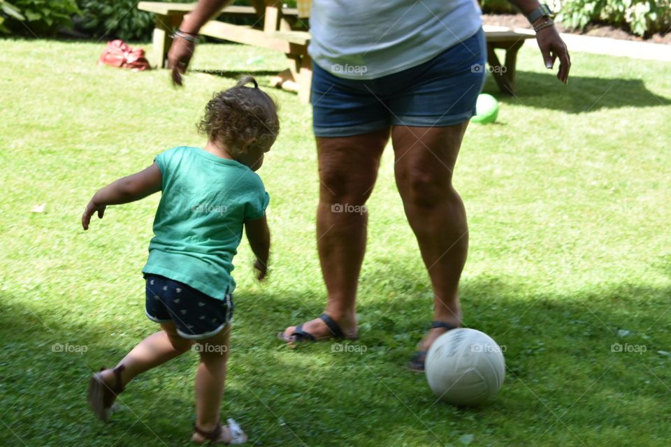 Adult and child in summer clothing kicking a soccer ball on a green lawn. 