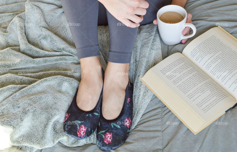 Woman's slippered feet on a bed next to an open book and holding a cup of coffee