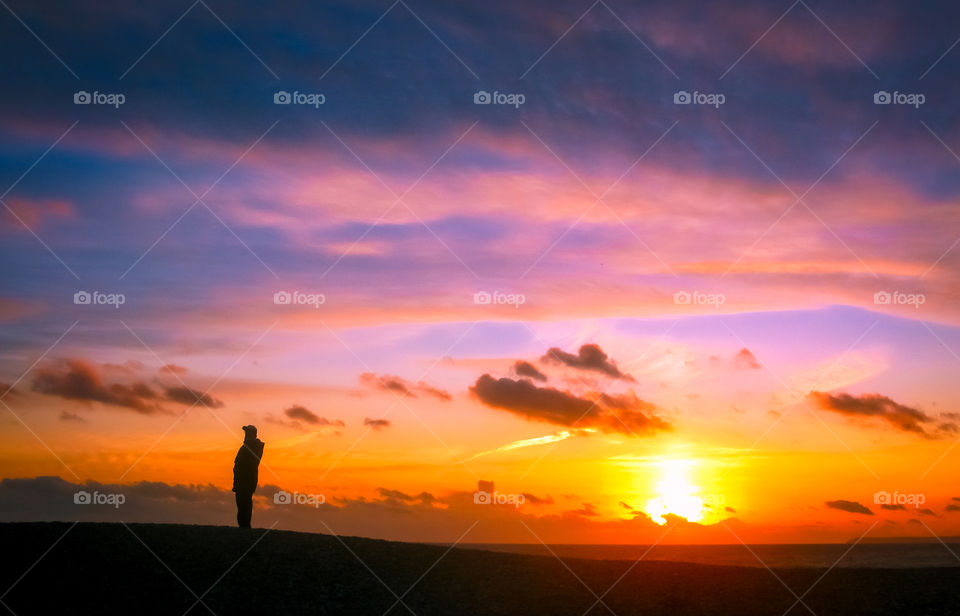 A man, wrapped up in warm clothes, pauses as he enjoys an evening stroll on the beach, he is silhouetted against a colourful wintery sunset