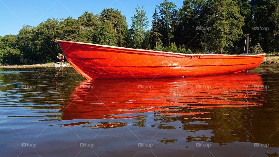 The red boat is parked on the water near the shore. Summer time. Reflection of a boat on the water