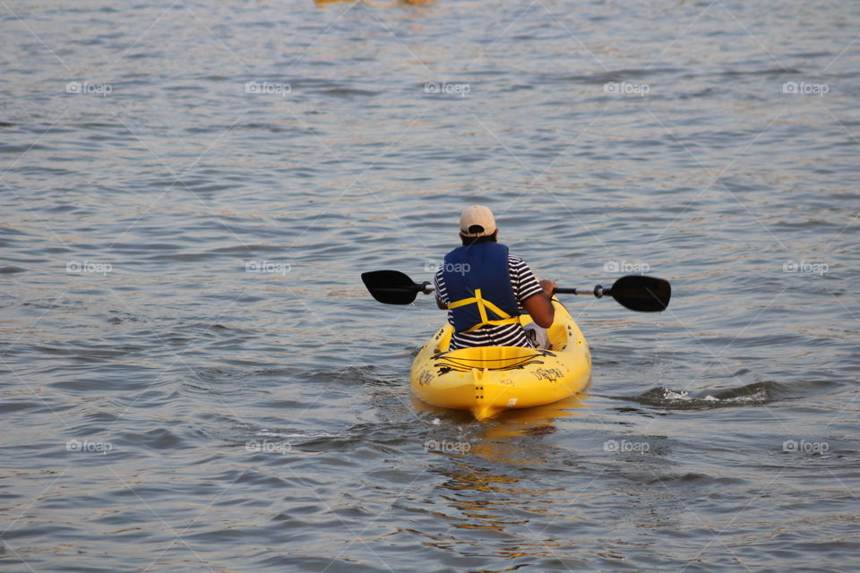 Man in blue life vest, striped shirt and beige baseball cap paddles bright yellow canoe on Hudson River on May evening 