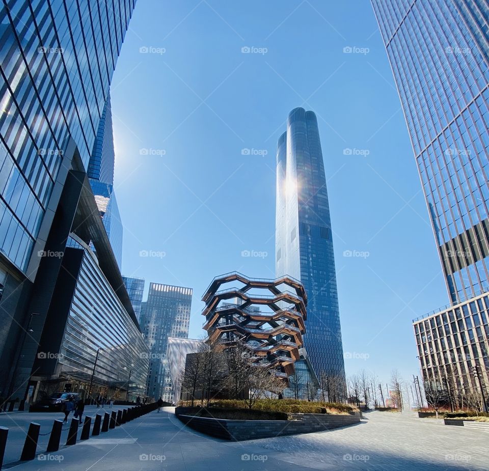 The Hudson Yards-Glass and steel-sleek skyscrapers with the iconic Vessel in the foreground and the sunny blue sky in the background.