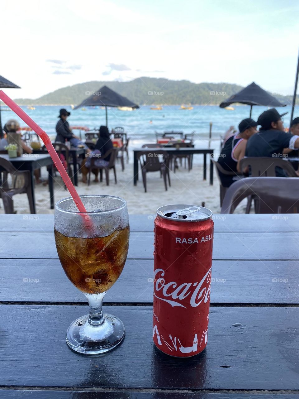A can of coca cola and a tall glass on a table with a sea view.