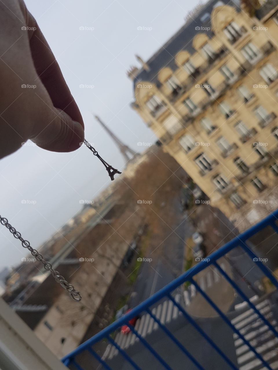 Hand holding a silver chain necklace with small charm of Eiffel tower from view of window balcony, same as real big Eiffel tower in Paris, France, Europe
