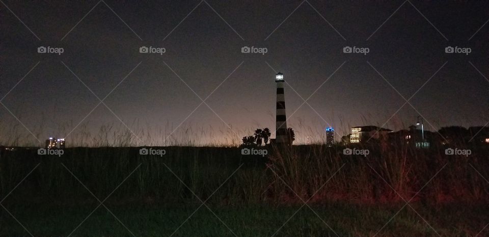 A lighthouse standing in a field at night time with some city buildings in the background.