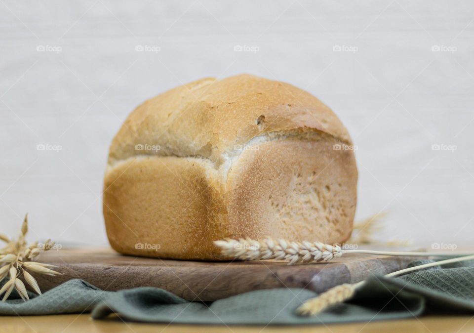 One square white wheat bread on a wooden board with ears and a cotton kitchen napkin lies on a table against a white blurred brick wall, close-up side view. The concept of baking bread.