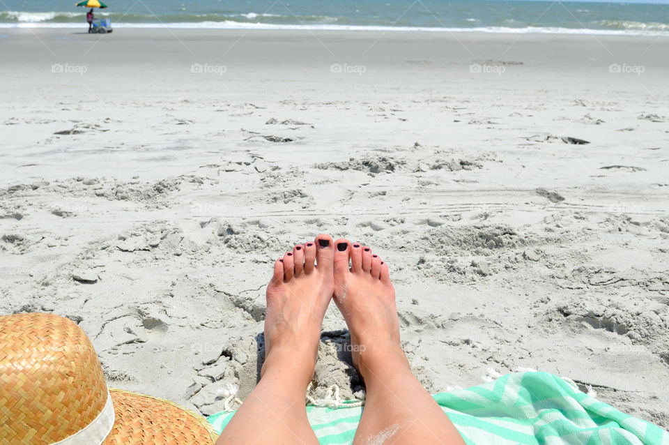 View of a woman's feet on a beach with a towel and hat and view of the ocean shore