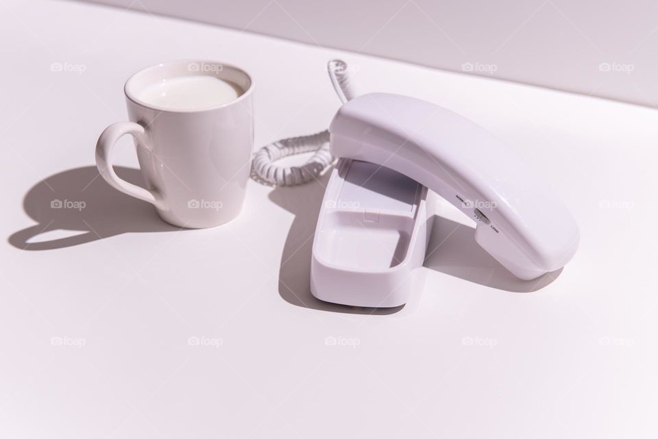 Hard light monochromatic minimalist image of a retro white corded telephone with a white mug of milk 