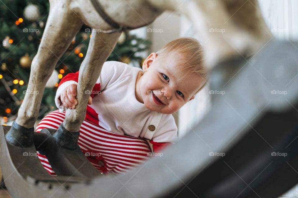 Little baby girl in red Santa costume looks out from behind toy wooden horse in room with Christmas tree at the  home