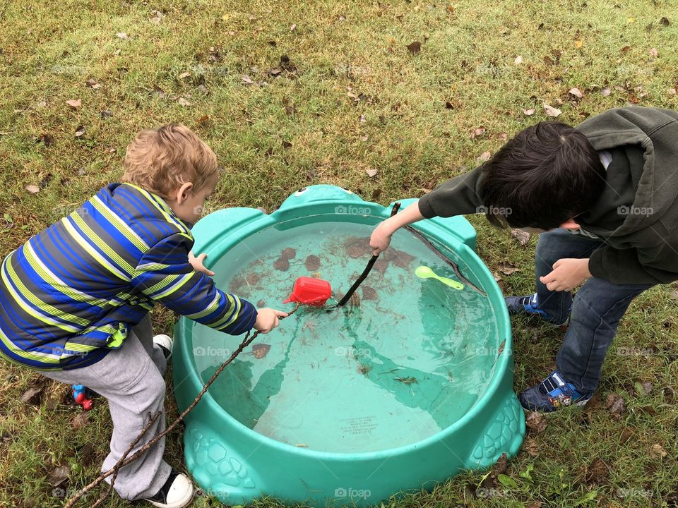 Rainwater filled pool with some dirt and a few fall leaves and two little boys with stick fishing poles playing fishing with their toys posing as fish. 