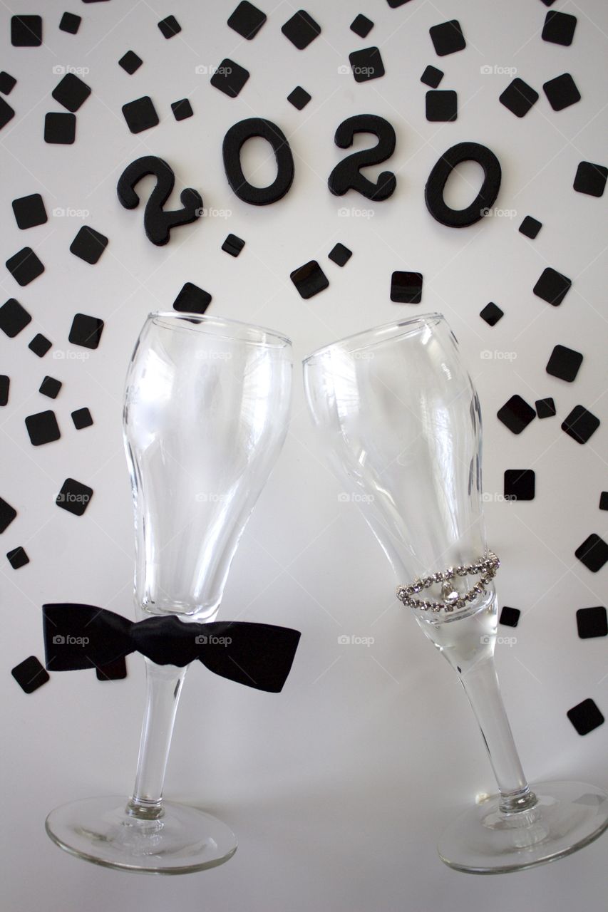 Conceptual overhead view of champagne glasses representing a man and a woman with the year ‘2020’ in black with black confetti on a white background - portrait format 