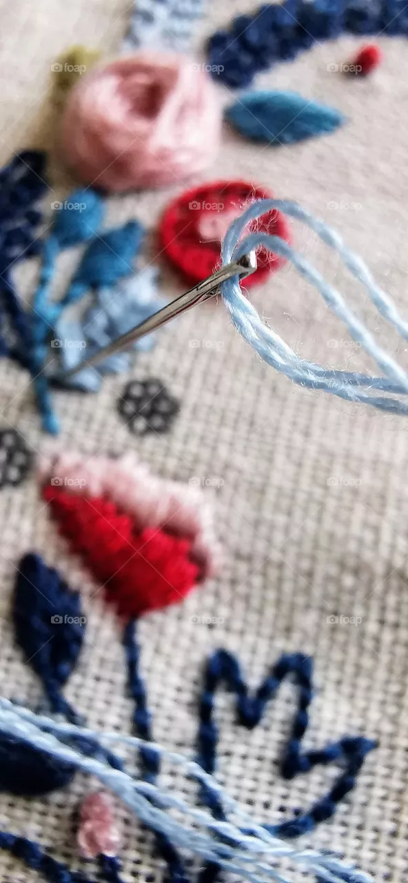 Needle with light blue thread in linen with embroidered blue, pink and red flowers and leaves in soft focus in the background