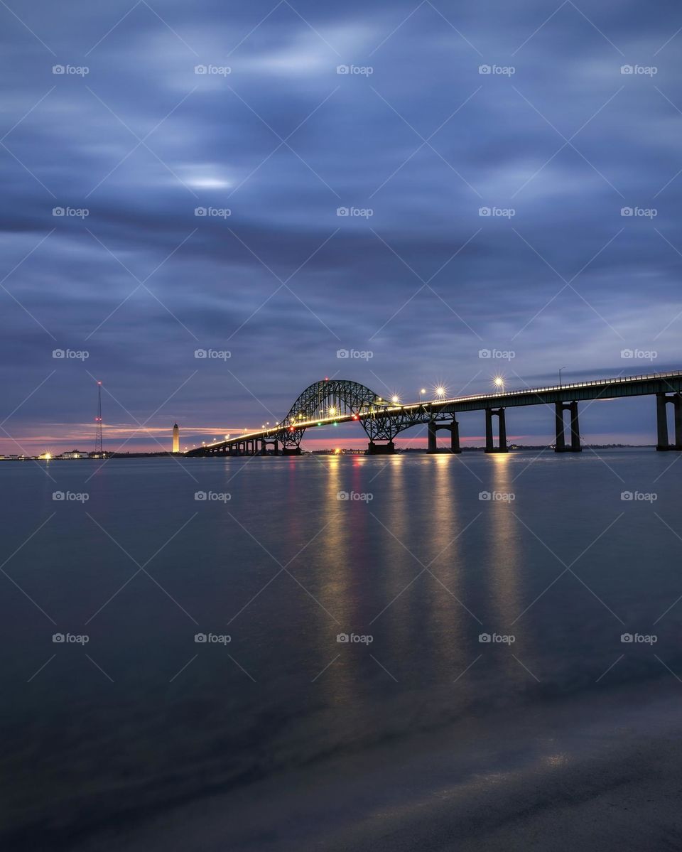Long steel tied arch bridge during blue hour with golden light from the street lamps reflecting in the water
