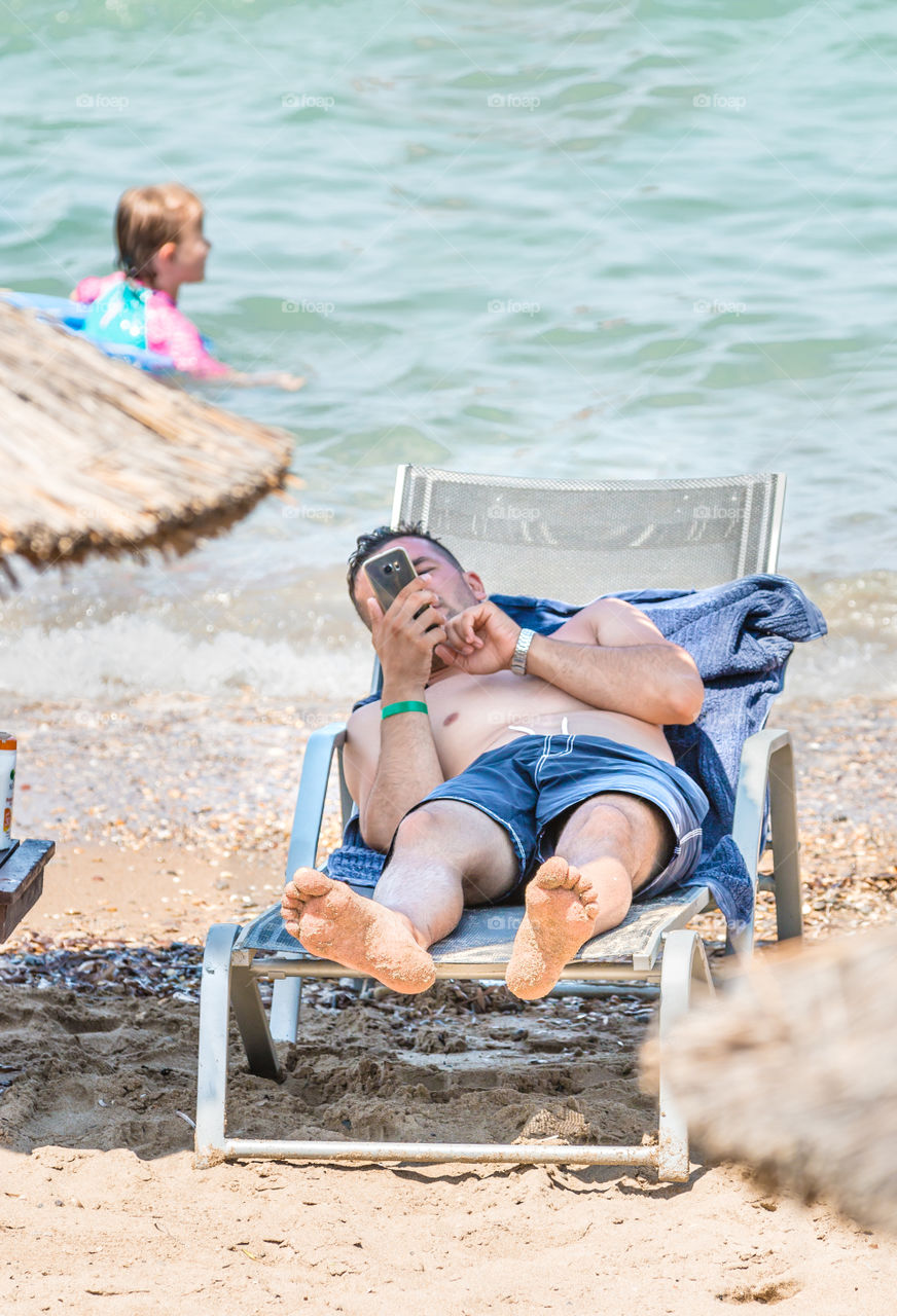 Man On Vacation Lying On A Sunbed And Using His Cellphone On The Beach
