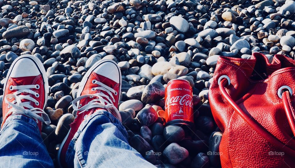 A can of Coca Cola on the beach with red sneakers and red leather bag.