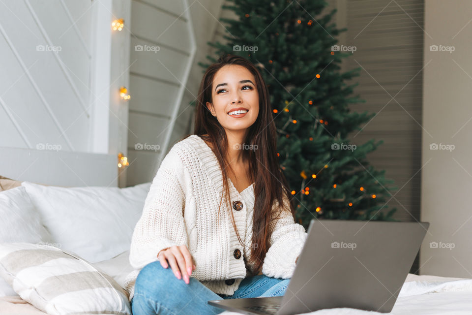 Young woman teenage girl in cardigan using laptop on bed in room with Christmas tree at home 