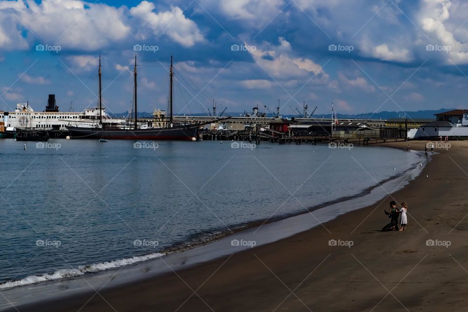 View of the beach at Aquatic Park in San Francisco with the skyline in the background and sailing ships on a beautiful cloudy day, father and daughter enjoying a beautiful day at the beach 