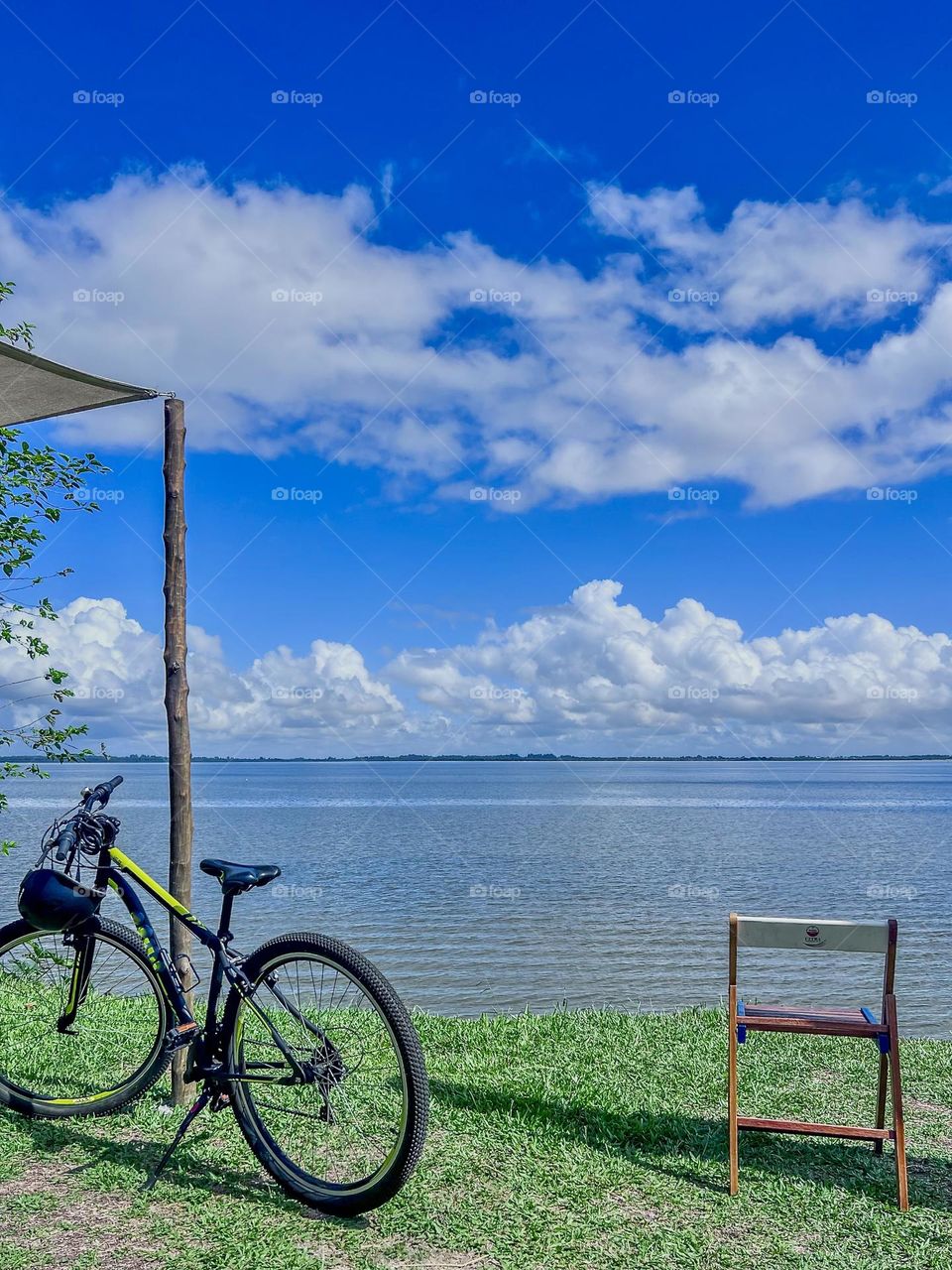 Parked next to a simple wooden chair on grassy banks, with a panoramic view of calm waters under a vibrant blue sky dotted with clouds. This image evokes feelings of relaxation and exploration, perfect for nature lovers.